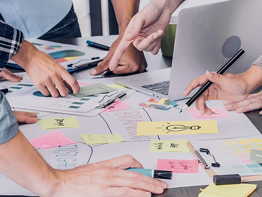 Image of memo notes and flip charts on a desk with a bunch of hands holding writing utensils.